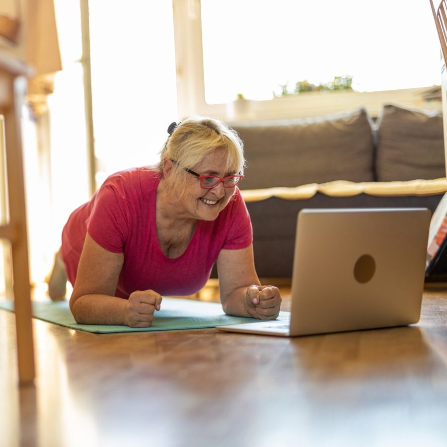 Woman smiling while doing a plank, looking at her laptop during a virtual coaching session