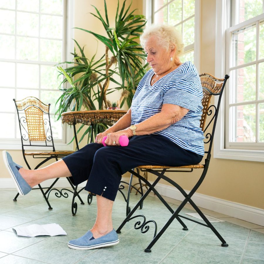 Woman in her 70s doing seated leg exercises with a hand weight in a sunny room